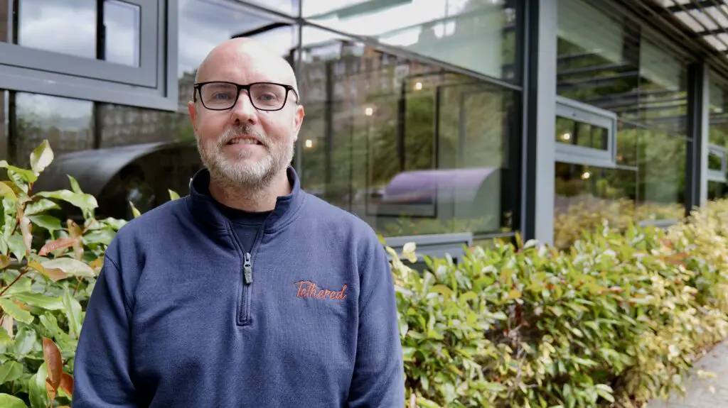 Tethered founder standing in front of green bushes.